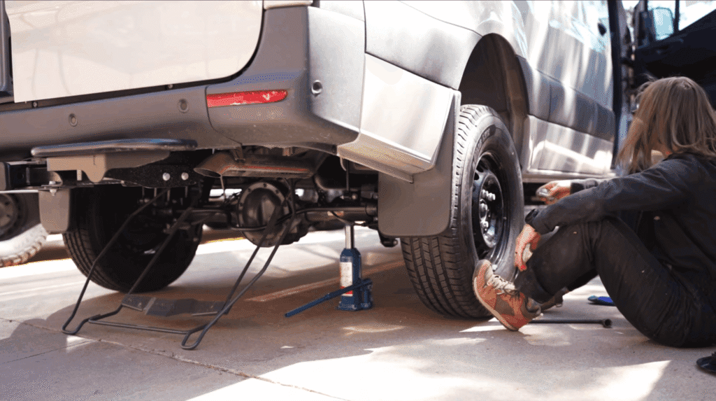 Hydraulic bottle jack positioned under the frame of a Mercedes Sprinter camper van with wheel removed
