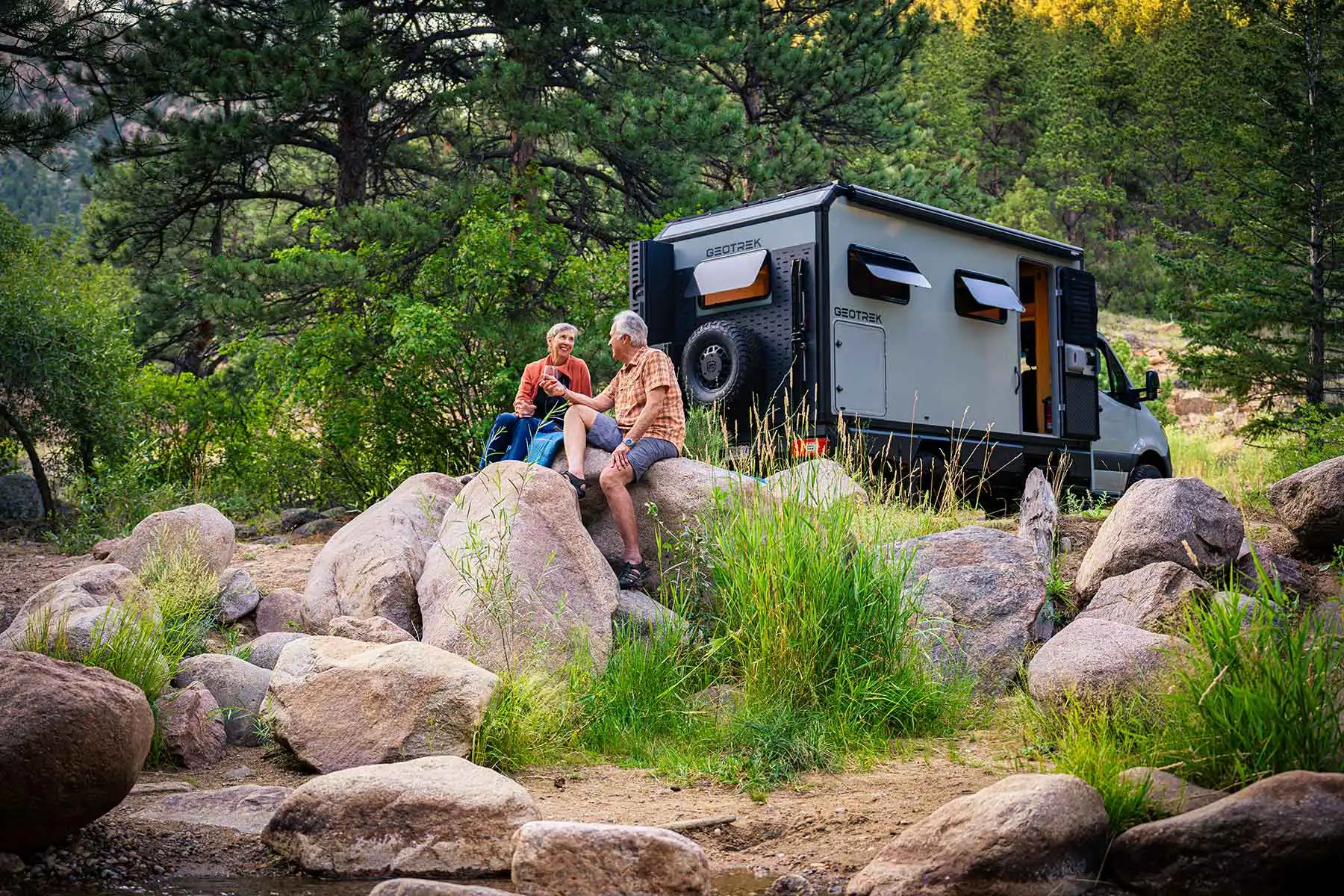 Couple sitting on streamside rocks next to a parked Geotrek Verus